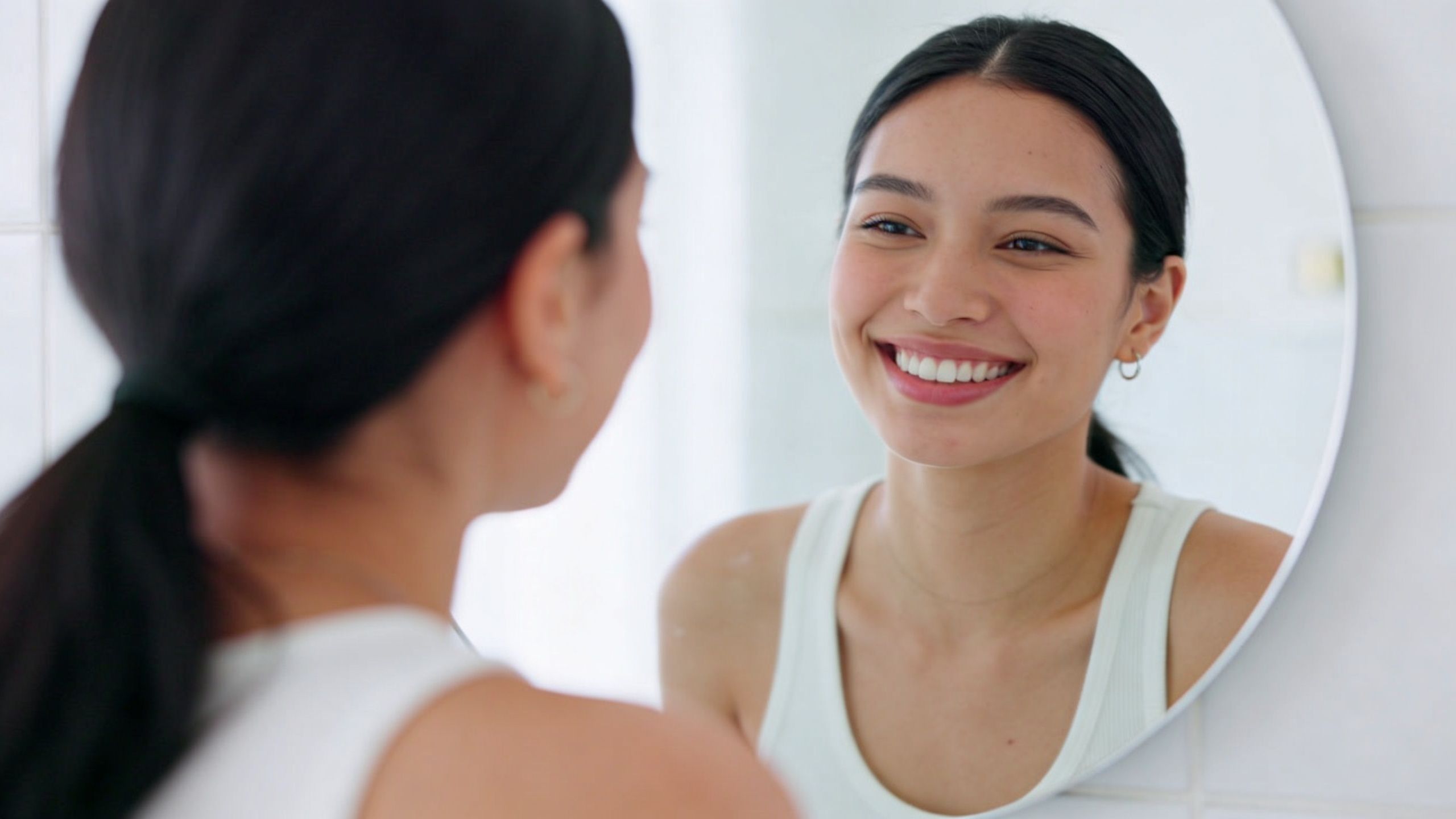 A woman with a bright, healthy smile looks at her own reflection in a round bathroom mirror.