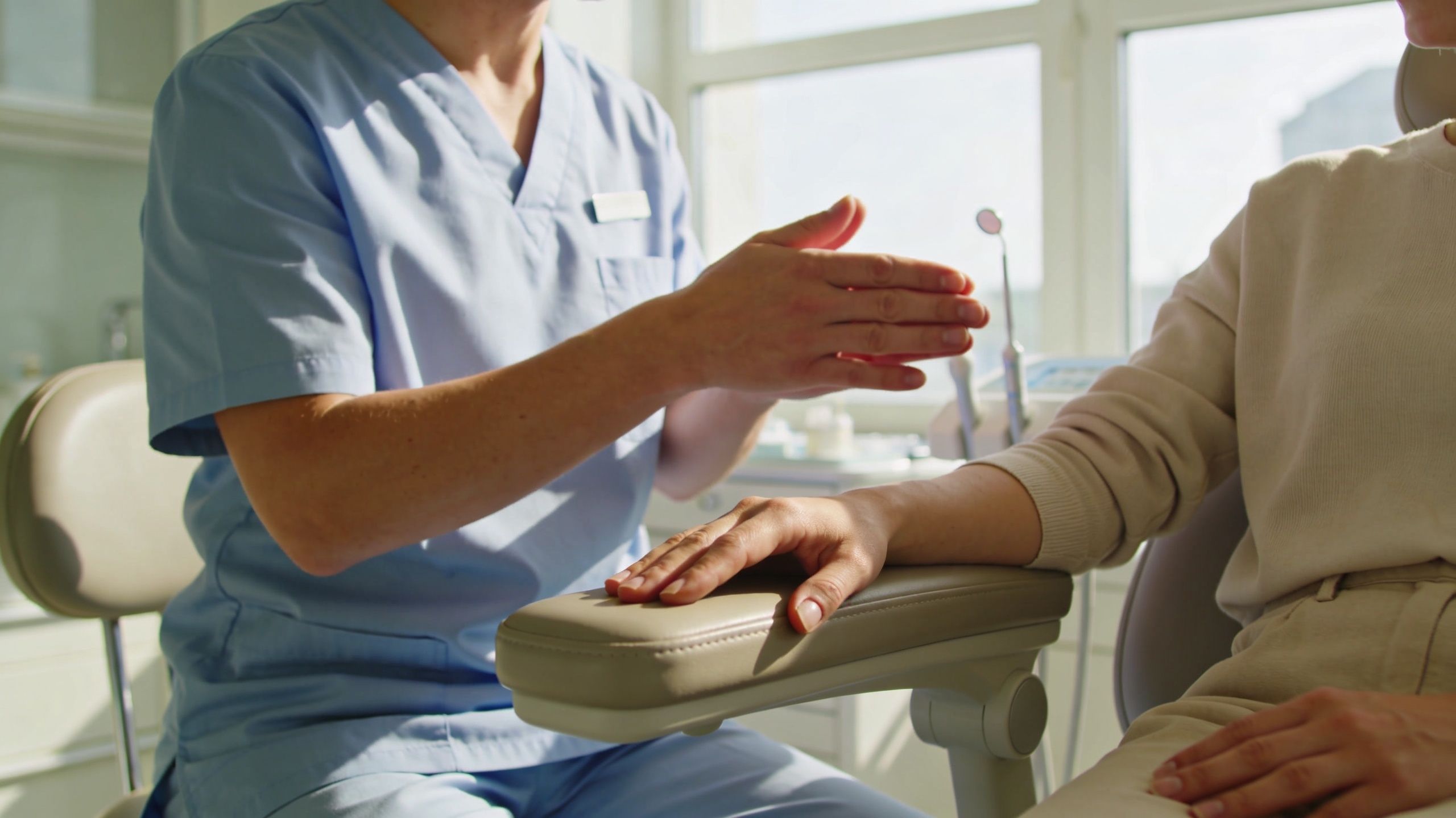 A dentist wearing blue scrubs gestures while consulting with a female patient in a bright dental office.