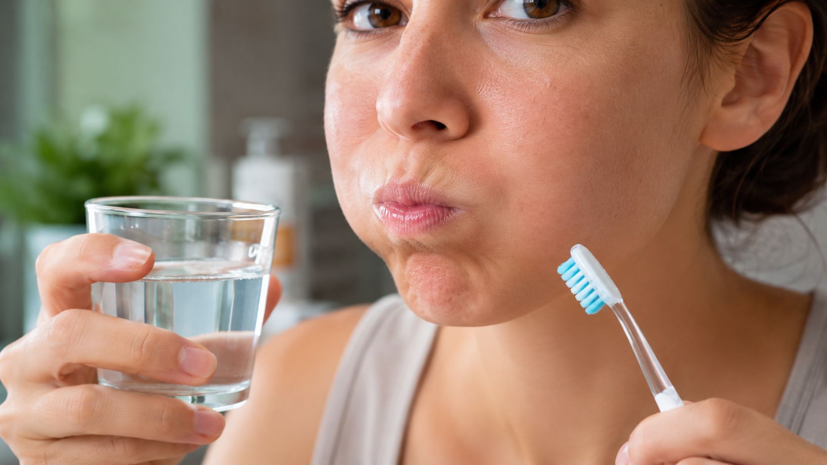 A woman holding a glass of water and a toothbrush while rinsing her mouth during daily hygiene.