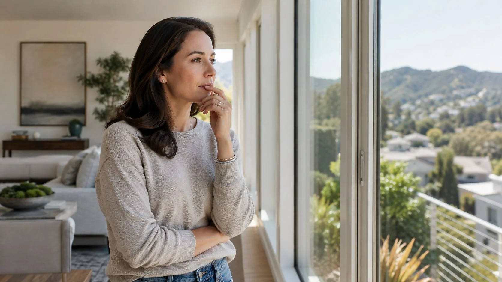 A thoughtful woman looking out of a large window with a scenic hilly neighborhood view in the background.