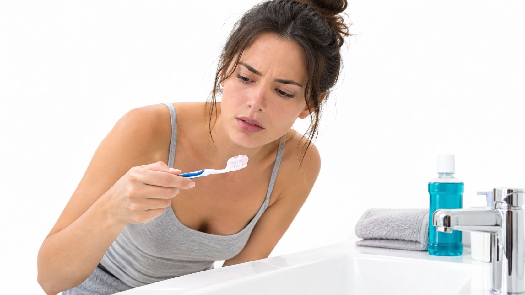 A concerned woman examining blood on her toothbrush while brushing her teeth in a bathroom setting.